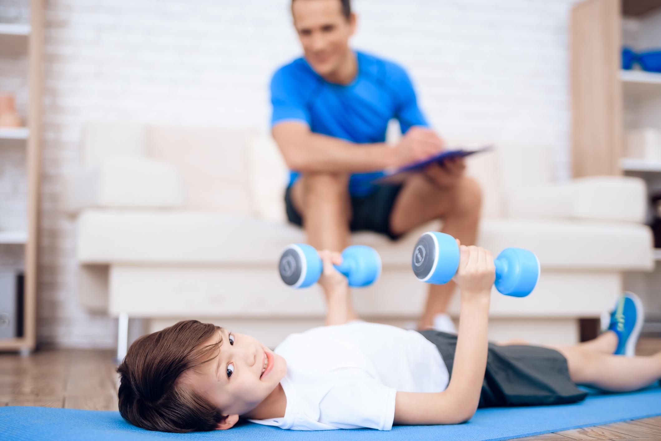 A boy is doing exercises with dumbbells on the floor. Next to his father, who trains a boy.
